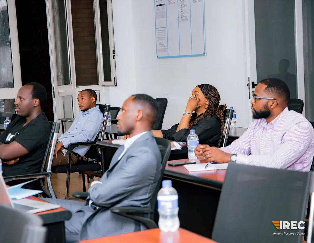 Participants seated at desks along one side of the room, listening and looking toward the front, with notebooks and water bottles in front of them.
