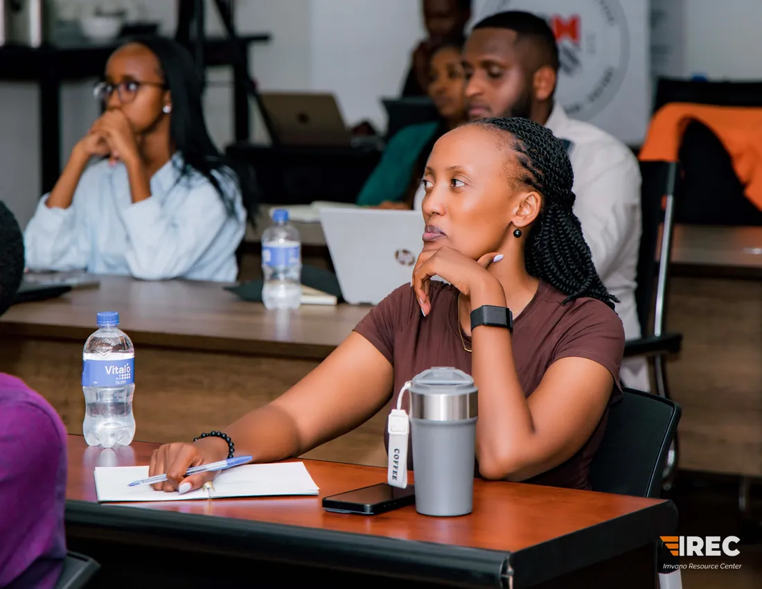 A participant resting their chin on a hand and listening, with a notebook, pen and travel mug on the desk and colleagues seated behind.