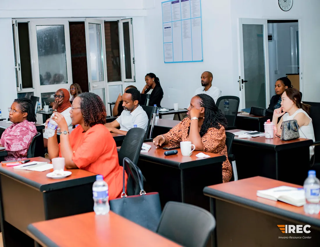 A bright classroom with participants at spaced desks facing the front, a wall chart and a clock on the walls.